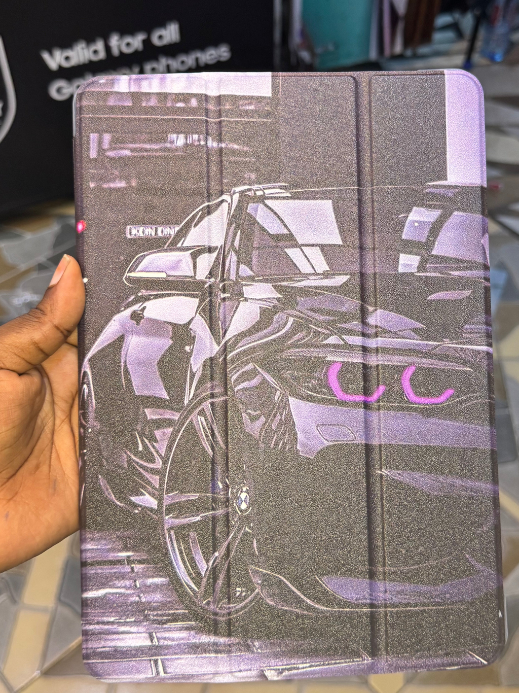 Hand holding a purple and gray notebook with a car design on a tiled floor background.